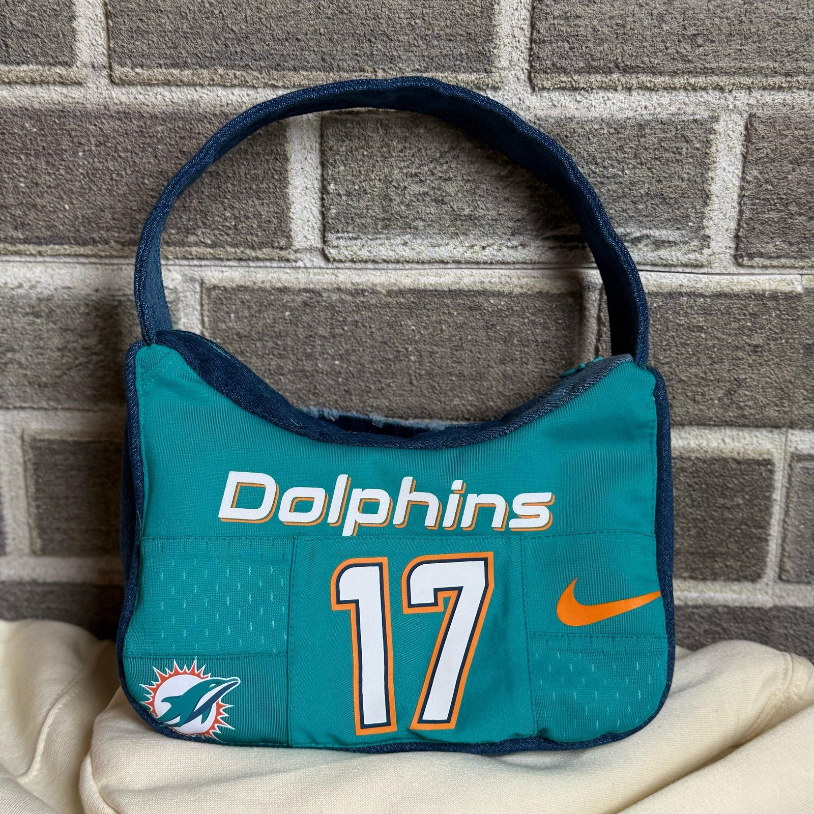 Upcycled denim shoulder bag with a Miami Dolphins patch and distressed detailing, displayed against a brick wall.