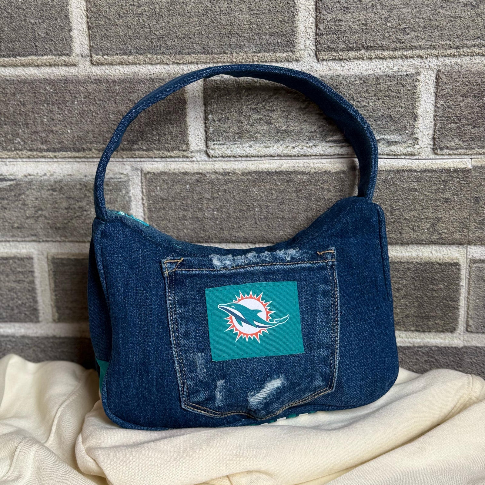 Upcycled denim shoulder bag with a Miami Dolphins patch and distressed detailing, displayed against a brick wall.